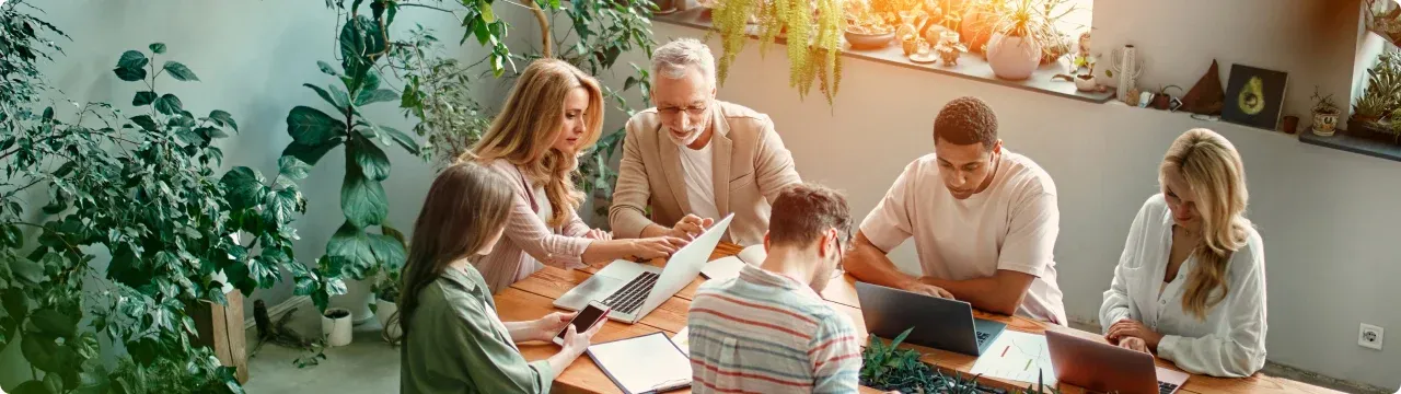 A team gathered around a table with laptops and notebooks in a plant-filled workspace.