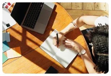 Person writing in a notebook beside a laptop on a wooden table.